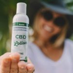 a woman wearing a black hat and a white top showing a white plastic bottle of CBD lotion on top of her palm