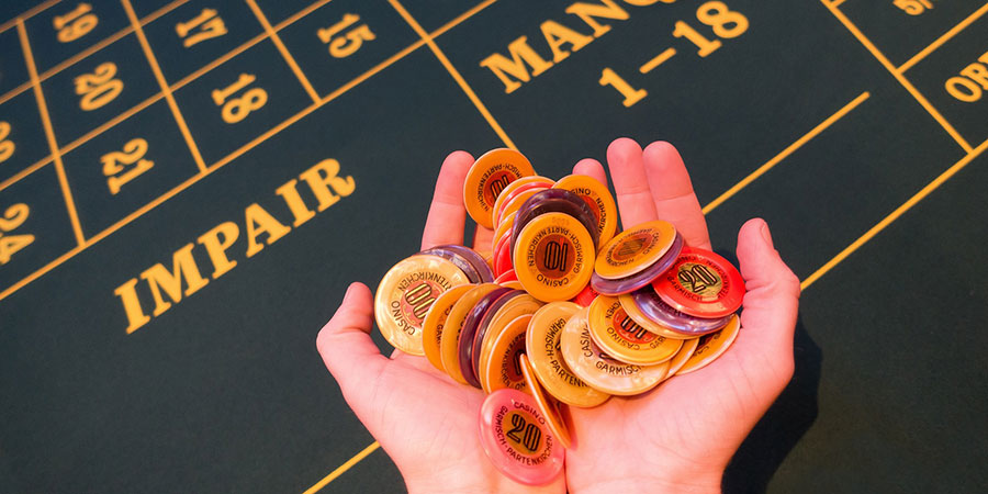 several casino chips on a person's hands and a roulette table in the background