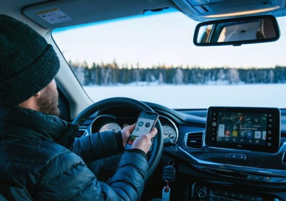 Driver in a midsize SUV holds a smartphone as if applying for auto financing, with car keys on the console, parked beside a snowy lakeside lined with boreal pines at golden hour.