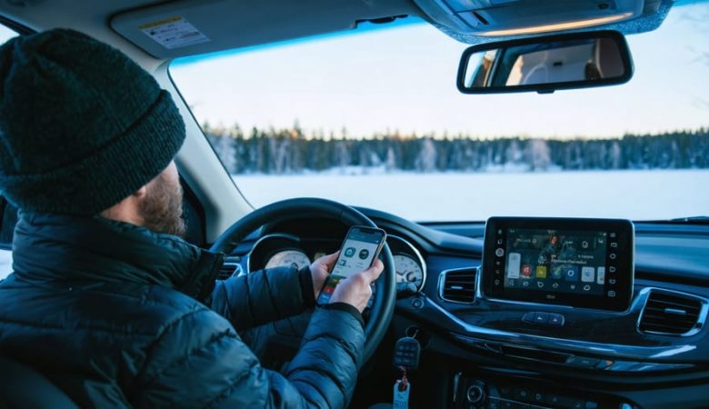 Driver in a midsize SUV holds a smartphone as if applying for auto financing, with car keys on the console, parked beside a snowy lakeside lined with boreal pines at golden hour.
